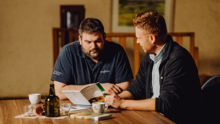 Zwei Männer sitzen am Tisch und besprechen Unterlagen bei Kaffee.