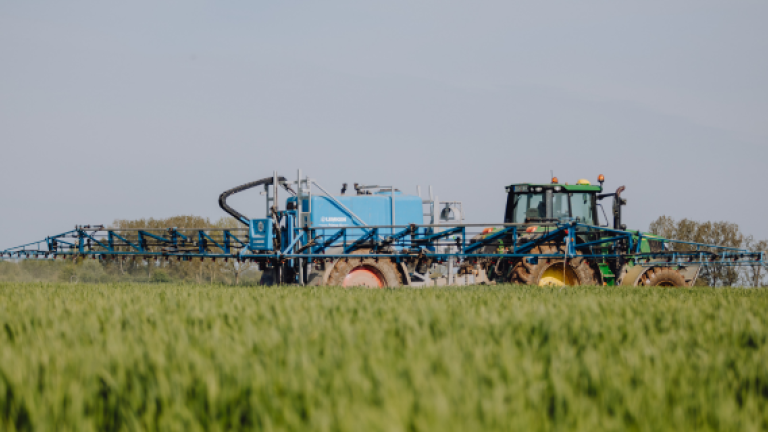 Blauer Traktor mit Anhänger auf Feld; im Vordergrund grünes Getreide, blauer Himmel.